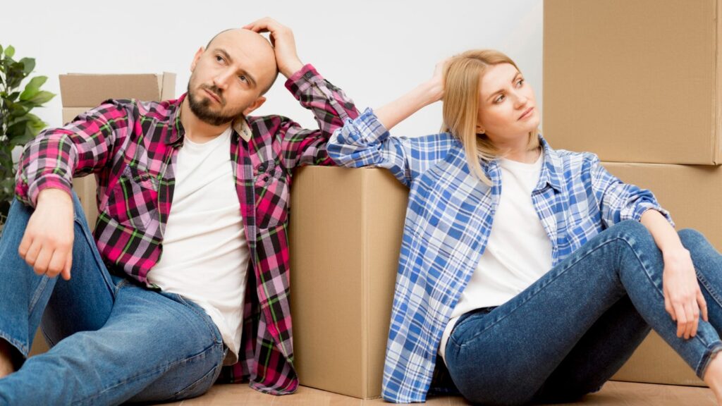 A couple sitting on the floor surrounded by cardboard moving boxes, looking stressed and uncertain about their home situation.
