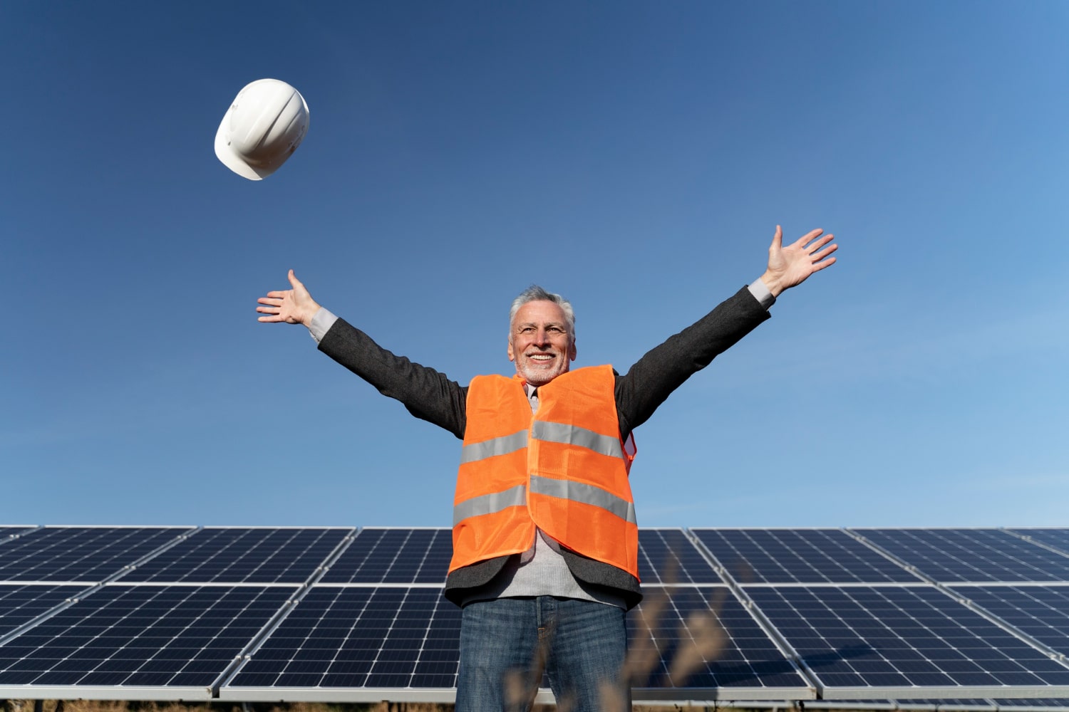 Smiling technician in safety vest celebrating in front of solar panels, tossing hard hat into the air.