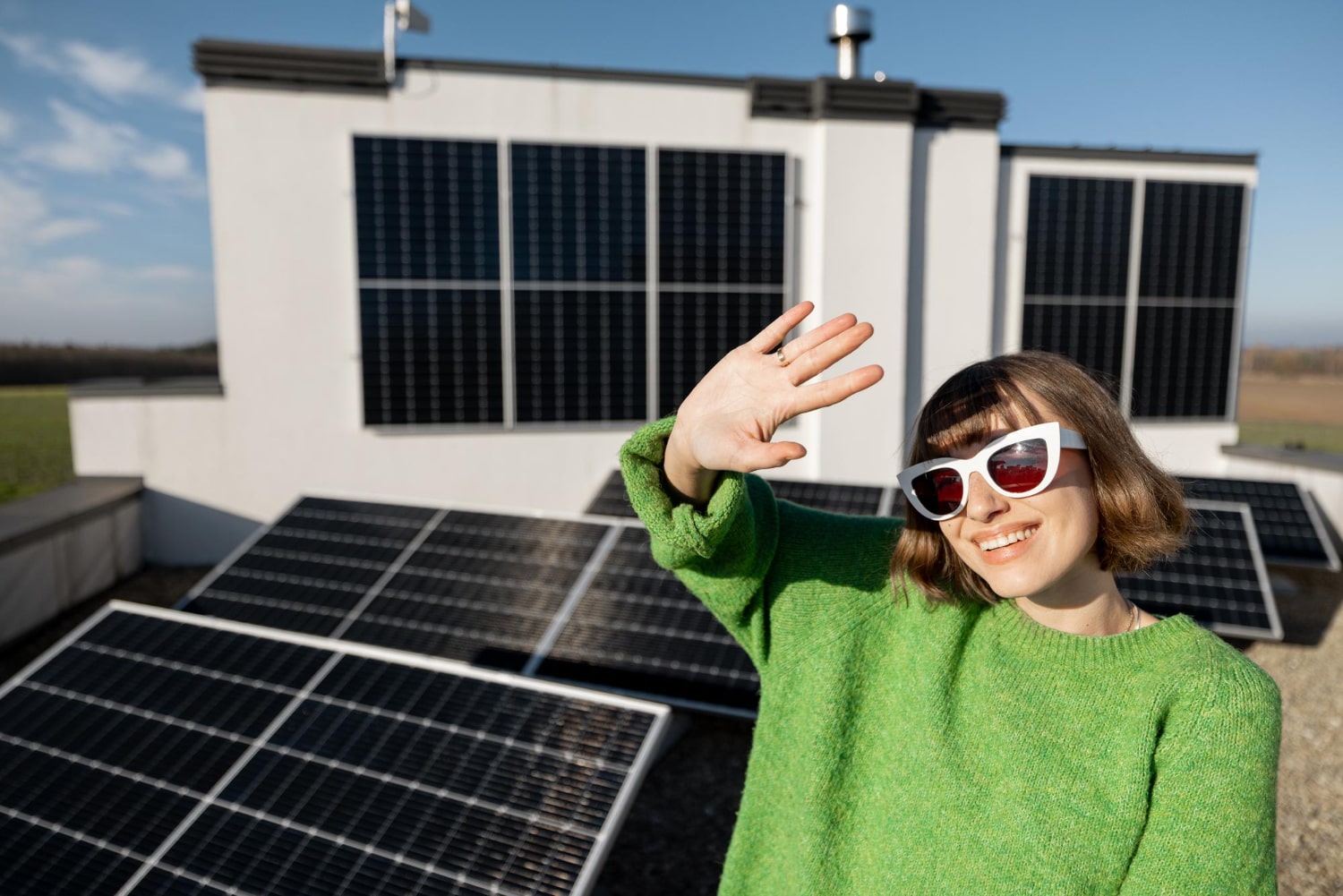 Smiling woman in sunglasses standing near rooftop solar panels, raising her hand in the sunlight.