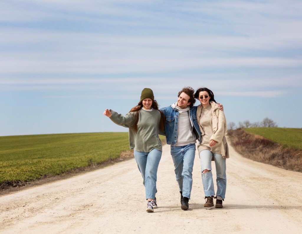 Three friends walking together on a rural dirt road surrounded by open fields under a bright sky.